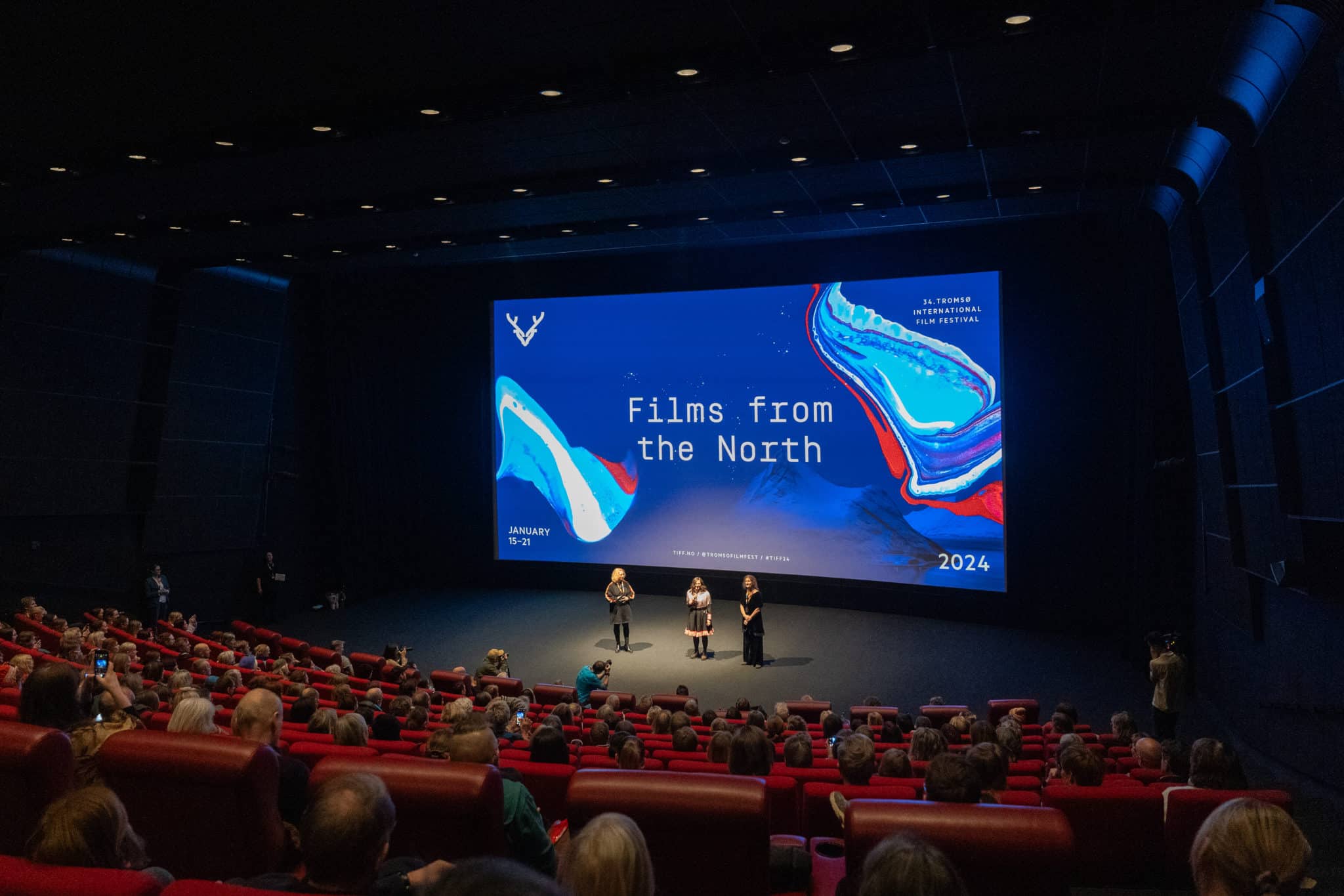 Three people standing in the spotlight in front of a blue cinema screen at TIFF 2024.