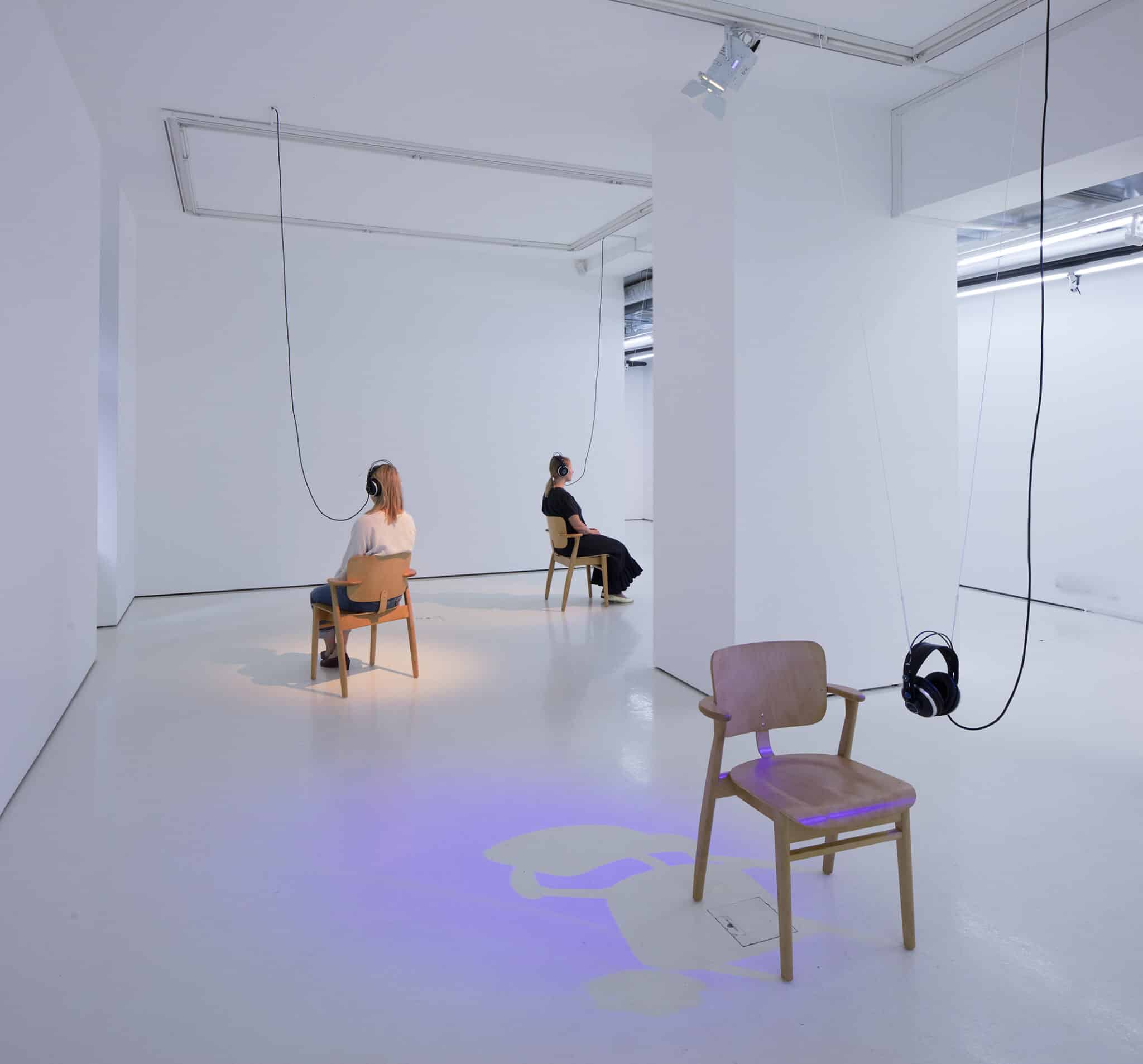Exhibition photo showing two people sitting on chairs in a white room, listening to a sound work through headphones.