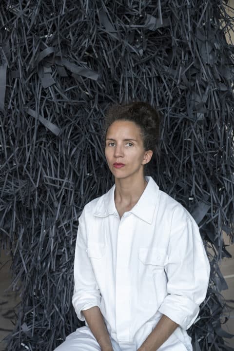 Portrait photo of a woman sitting on a chair in front of a black background. The woman is dressed in a white overall.