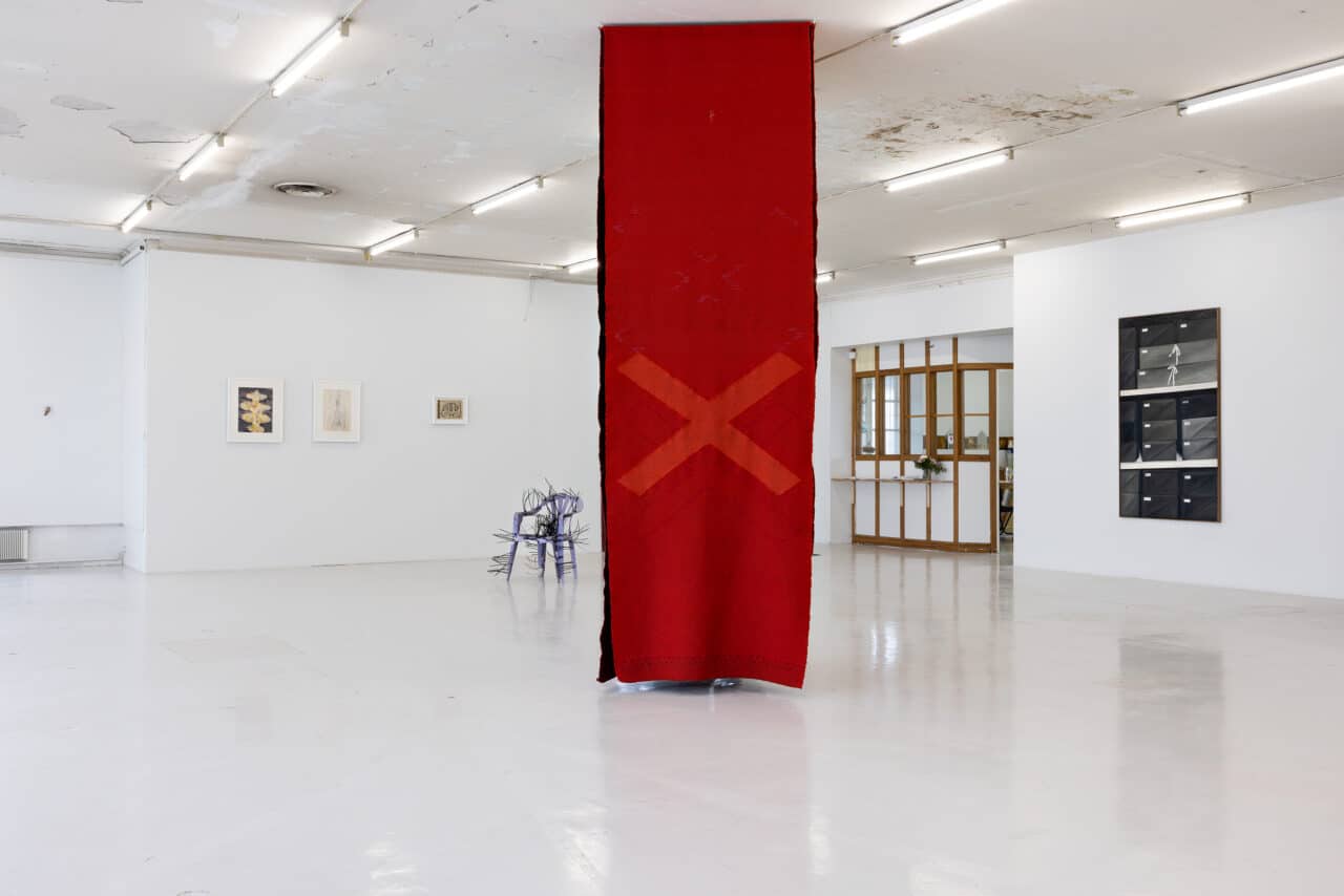 Tapestries—woven in varying tones of red and adorned with the form of a cross in a light-coloured gallery space. Behind and on the sides of the tapestries are other artworks, such as a thermoplastic chair sculpture stitched together with cable ties.