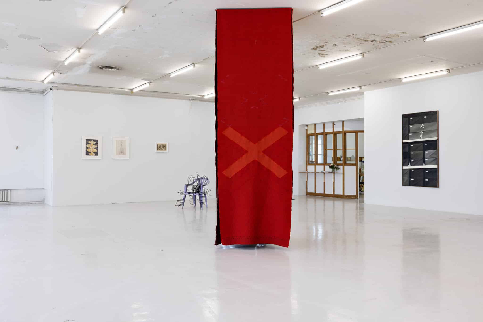 Tapestries—woven in varying tones of red and adorned with the form of a cross in a light-coloured gallery space. Behind and on the sides of the tapestries are other artworks, such as a thermoplastic chair sculpture stitched together with cable ties.
