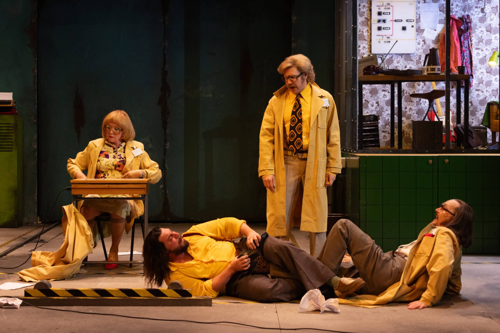Scene photo from a theatre play: two men are lying on the ground, one holding a shoe and the other looking surprised, while a person stands and shouts at them, and a woman sits at a table. All four characters are dressed in yellow, wearing large, retro-style glasses and clothing.