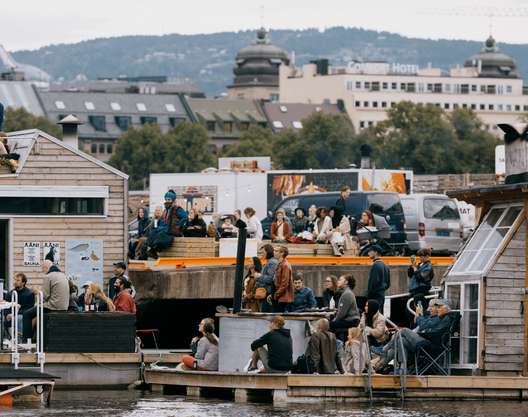 People sitting at a bridge by the water enjoying sound Sauna concert series at Oslo Badstuforening during a warm season.