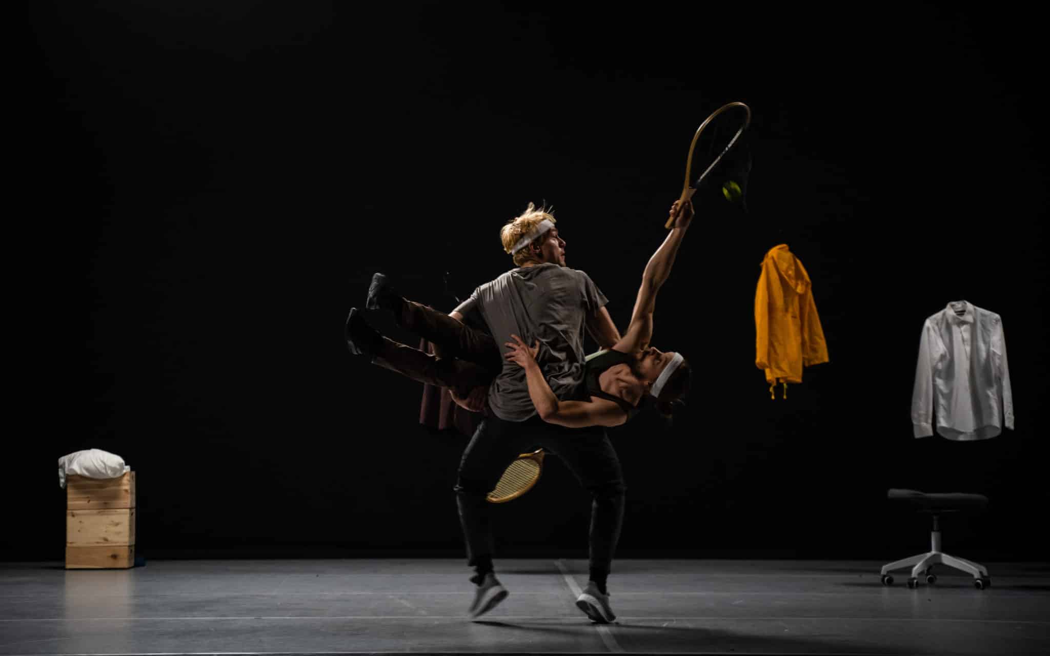 Still image from the performance. Dimly lit stage, where two performers are in the middle of an acrobatic stunt, with tennis rackets and a ball. A jacket, shirt and seatings can be seen in the background of the stage.