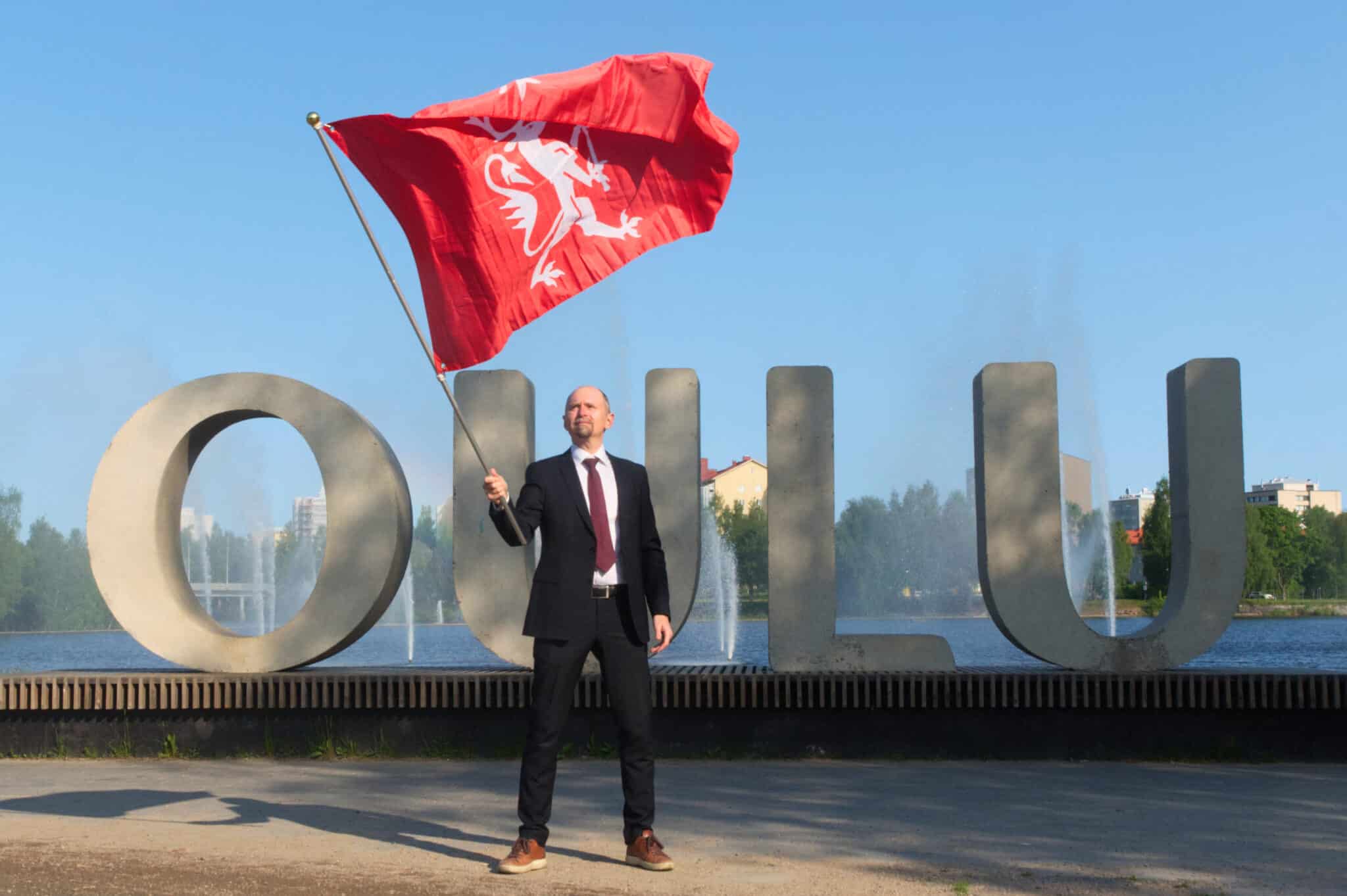 A person in a suit holding a flag in front of a sign reading: Oulu