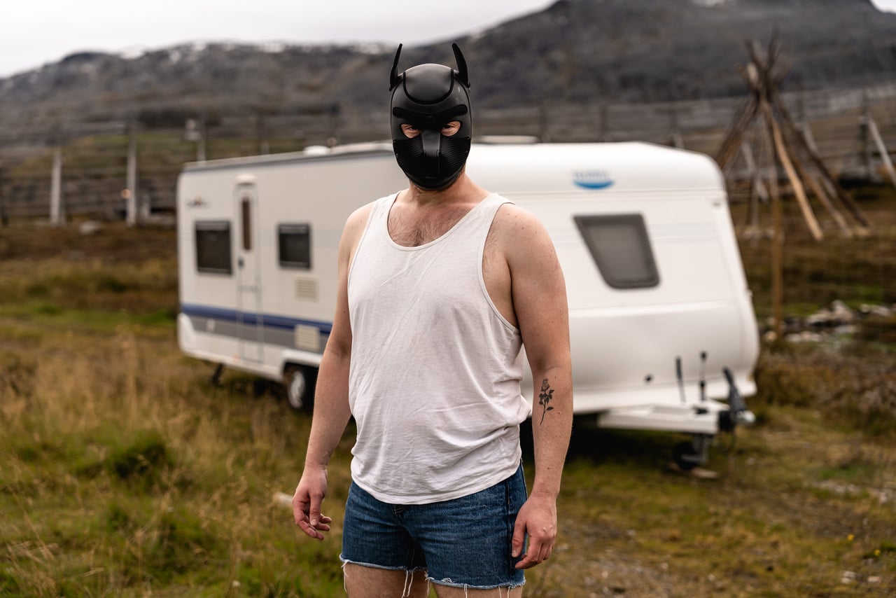 A standing person with a tank top and jean shorts stares at the camera. Their face is covered with a black mask with ears. Barren landscape with a white trailer in the background.