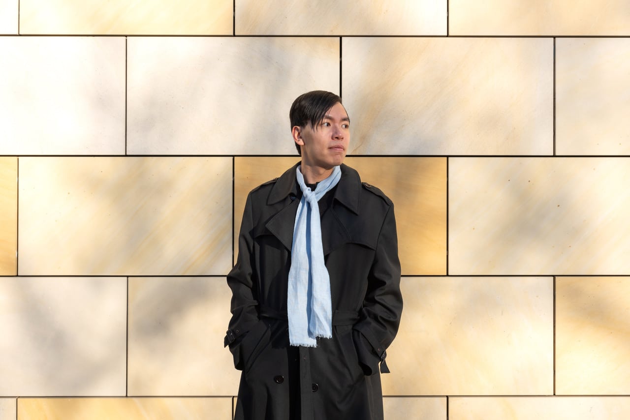 The composer Tze Yeung Ho is posing outside in front of a colorful stone wall in daylight.