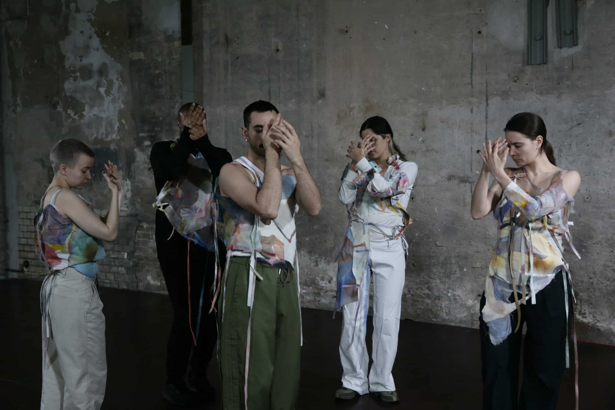 Five dancers in front of a concrete wall covering their closed eyes with their hands.