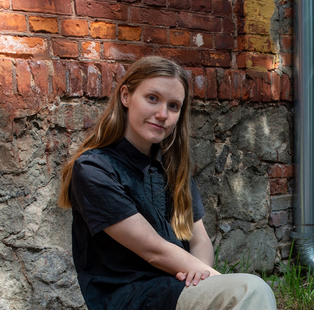 Nærbilde av en kvinne med langt hår iført en svart skjorte, sittende mot en murvegg.