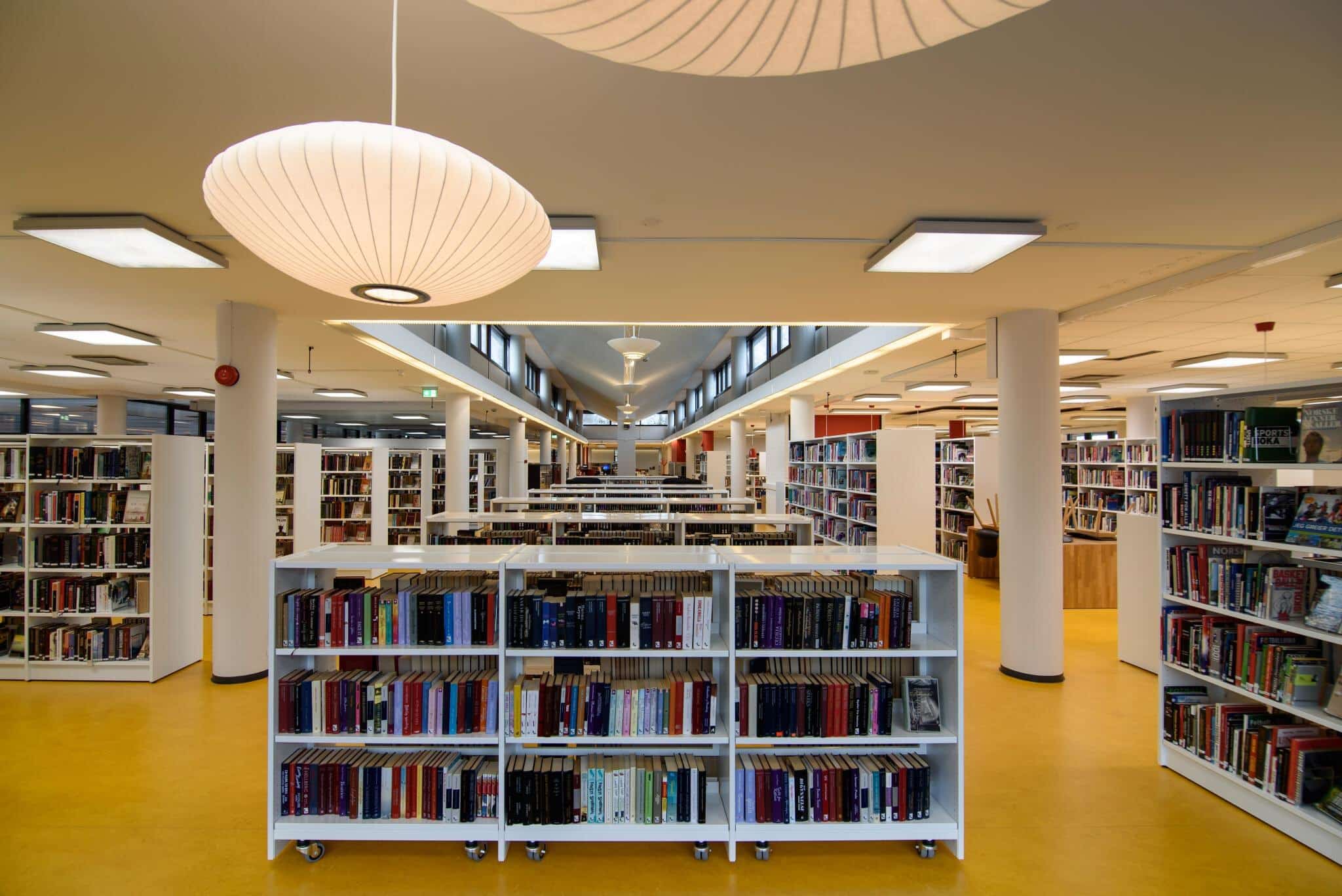 Photo: Inside of a library. The floors is yellow and there are many white colored bookshelves.