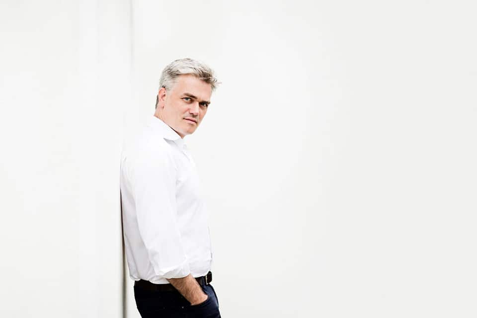 A medium close-up of a man standing and leaning to a white wall.