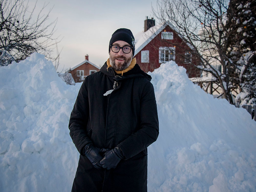 A photo of a man standing in front of pile of snow. There is a read house in the background.