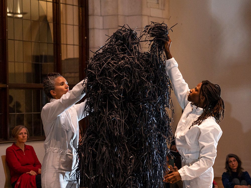 Art performance where two persons dressed in white overalls are putting shredded black paper on a stand.