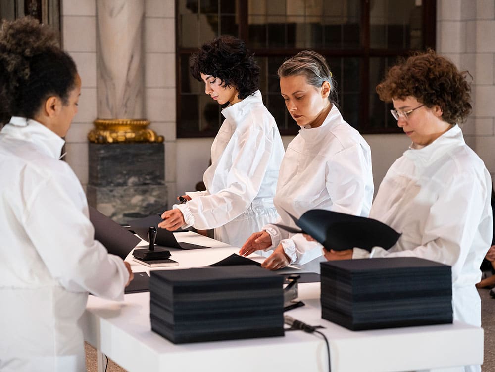 Four persons in white overalls are standing by a table handling black paper.