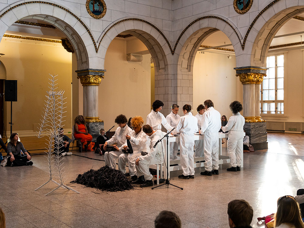 People in white overalls are standing around a white table. Three people with white overalls are sitting at the end of the table shredding black paper.