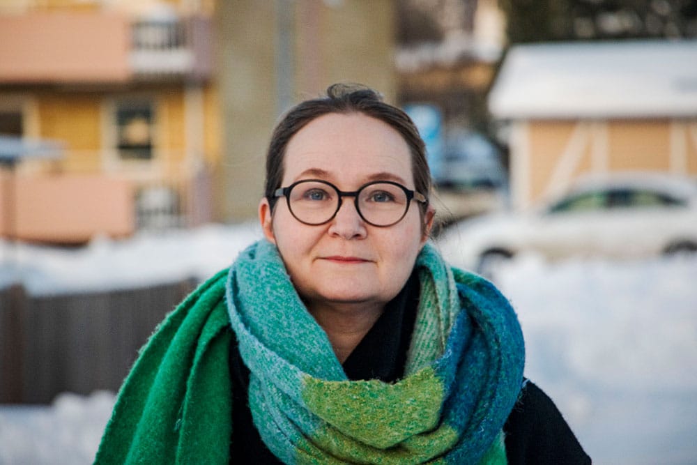 A close up photo of a woman. In the background there is snow and yellow houses.