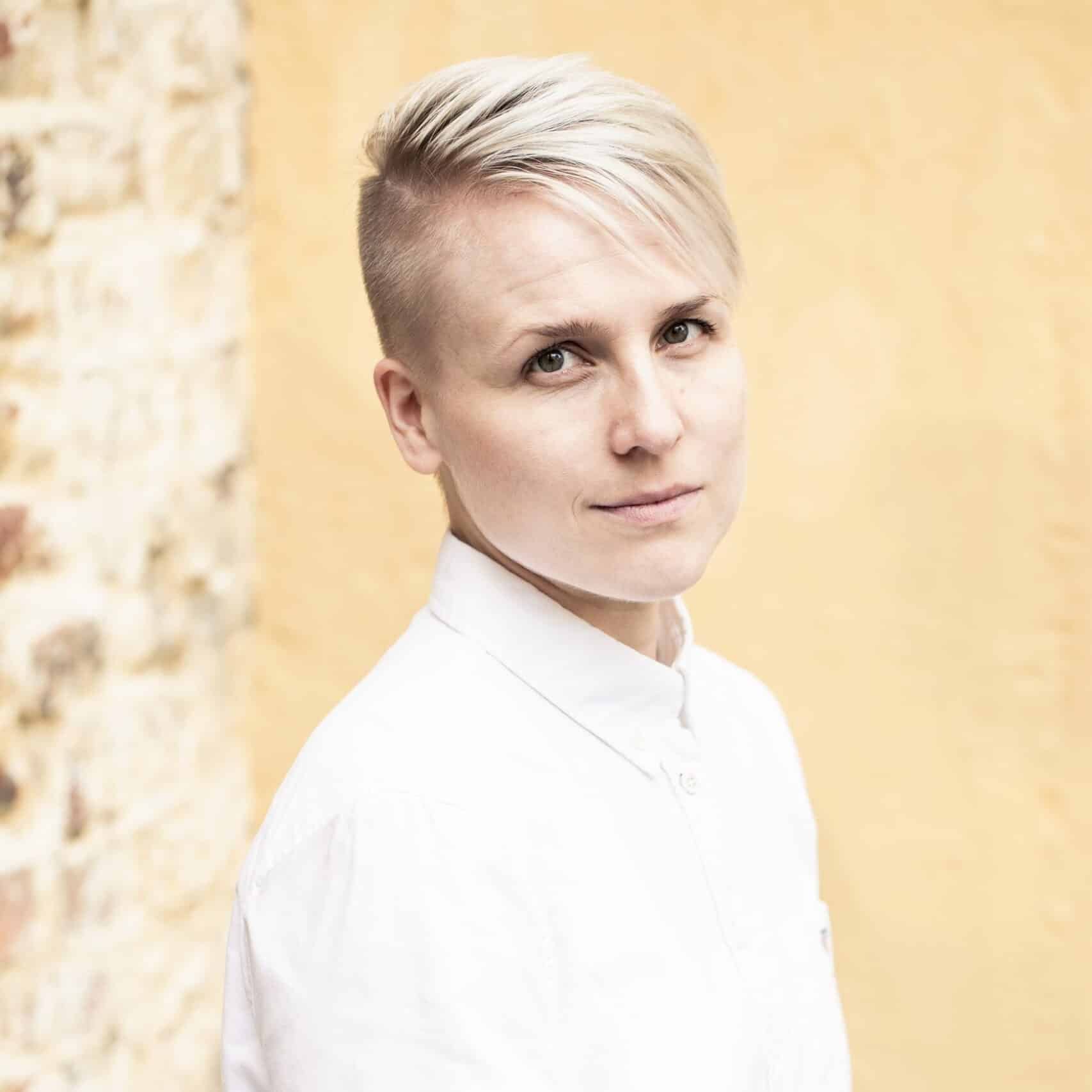 Portrait of Tiina Tuppurainen with short blonde hair and a white shirt on front of a yellow wall.
